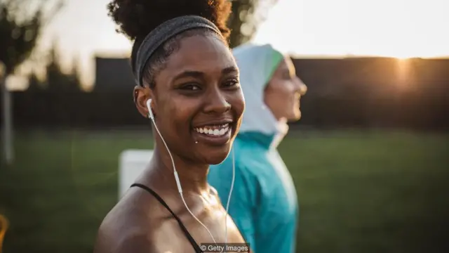 Mulher com traje esportivo sorri, e outra mulher, com véu na cabeça, aparece sorrindo no planoarbety sinaisfundo