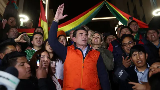 Rodrigo Paz, vestindo um colete vermelho e cercado por apoiadores, levanta o braço direito.