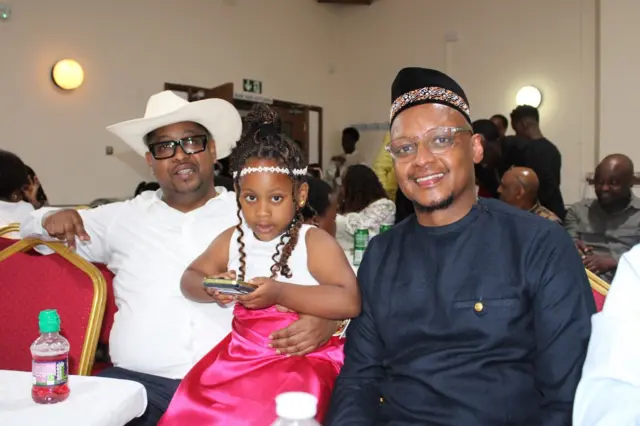 Umwana ufite telefone, yicaye hagati y'abagabo babiri