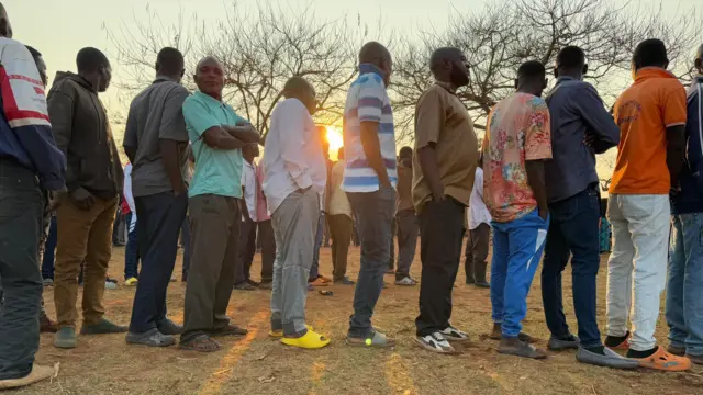 One wide shot from di side of queue of pipo wey dey wait to vote on a dusty, dry field. 