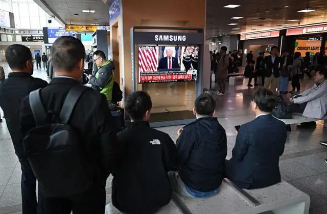 Transeúntes sentados en una banca de una estación de metro de Seúl, Corea del Sur, observan un televisor donde se transmite el discurso del presidente Trump