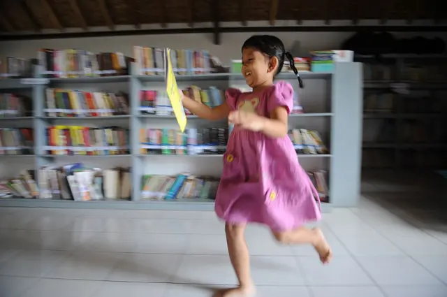 Seorang anak perempuan sedang berlari membawa buku di sebuah perpustakaan di daerah Bogor.