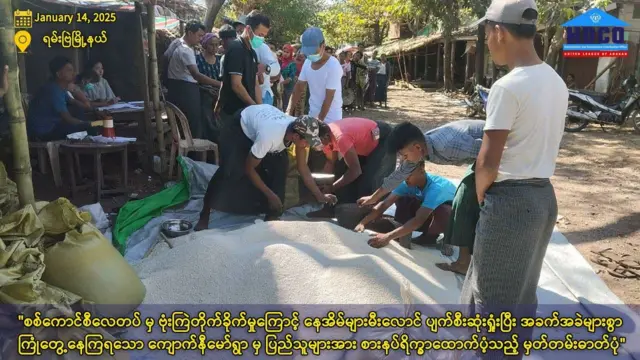 HDCO အဖွဲ့၏ ‌ထောက်ပံ့မှု မြင်ကွင်းလို့ဆို။ အမျိုးသားတစ်ချို့ မြေပေါ် အခင်းခင်းထားတဲ့ ဆံပုံပေါ်ကနေ ဆန်သဲ့ယူနေကြပြီး အနောက်မှာ လူတန်းရှည်ကို မြင်တွေ့ရ 