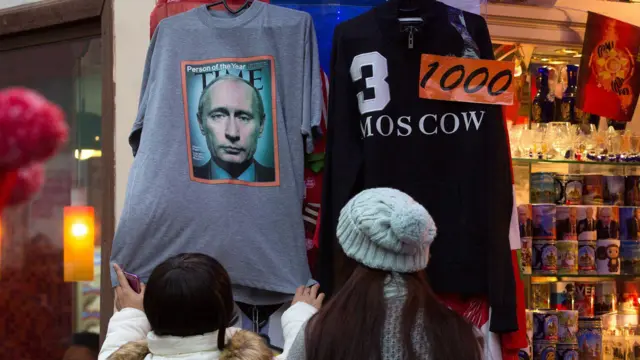 Duas mulheres usando chapéus examinam uma camiseta cinza com a capa da revista Time estampada, que está pendurada na porta de uma loja de souvenirs.