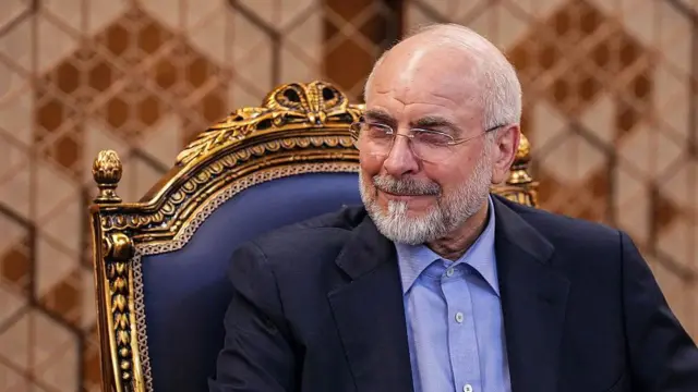 محمدباقر قالیباف، رئیس مجلس ایران روی یک صندلی با چوب طلایی نشسته است و لبخند به لب دارد.