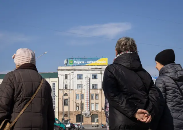 Три жінки середнього віку у верхньому одязі, які стоять на площі спиною до камери. Перед ними – світла багатоповерхова будівля з великим термометром і годинником на фасаді. На даху будівлі розміщений великий банер із текстом українською: "Харків – місто героїв!". Фон – ясне синє небо, безхмарна погода.
