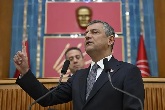 Özgür Özel kürsüde işaret parmağını kaldırmış, geri planda Atatürk büstü ve CHP'nin okları odak dışında görünüyor. 