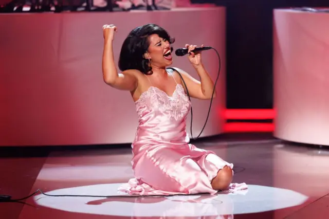 Raye sits on stage in a pink satin dress, singing into a microphone with her arm reaching out above her.