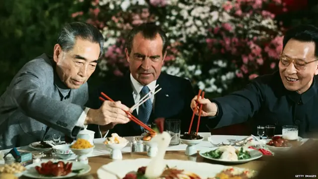 ਰਾਸ਼ਟਰਪਤੀ ਨਿਕਸਨ ਖਾਣੇ ਦੀ ਮੇਜ਼ ਉੱਤੇ ਚਾਪ ਸਟਿਕ ਫੜ ਕੇ ਤਿਆਰ ਬੈਠੇ ਹਨ ਜਦਕਿ ਉਨ੍ਹਾਂ ਦੇ ਨਾਲ ਸੱਜੇ ਹੱਥ ਚੀਨੀ ਪ੍ਰਧਾਨ ਮੰਤਰੀ ਜ਼ੌਏ ਇਨਲੈਅ ਅਤੇ ਸ਼ਿੰਘਾਈ ਕਮਿਊਨਿਸਟ ਪਾਰਟੀ ਦੇ ਆਗੂ ਚੈਂਗ ਚੁਨ-ਚਿਆਹੋ ਨਿਵਾਲੇ ਚੁੱਕਦੇ ਹੋਏ।