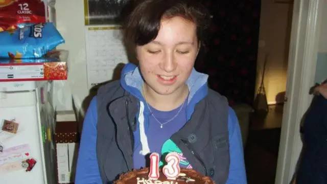 Gabby, üzerinde 13 rakamı bulunan çikolatalı doğum günü pastasına bakıyor. Mor bir bluz ve lacivert bir yelek giymiş.