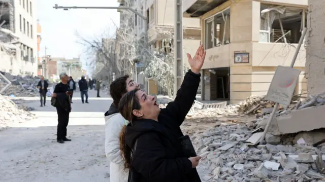 Жена и дечак гледају и показују руком око њих су разрушене зграде