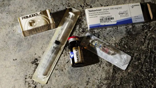 A imagem mostra diferentes itens associados ao uso de esteroides anabolizantes e testosterona dispostos sobre uma superfície de concreto. Entre eles, há duas caixas de medicamentos — uma identificada como Primobol-100 e outra como Testoviron Depot 100 mg/ml —, um frasco de vidro com tampa vermelha e duas seringas embaladas individualmente em plástico estéril. A composição destaca os produtos e os materiais utilizados para aplicação injetável dos hormônios.