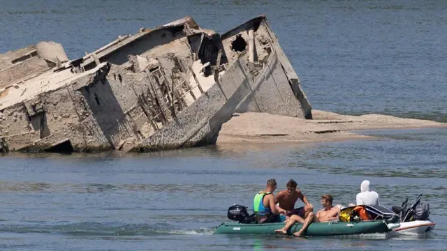 Destroços de um navio em um banco de areia, com pessoas observando em um barco