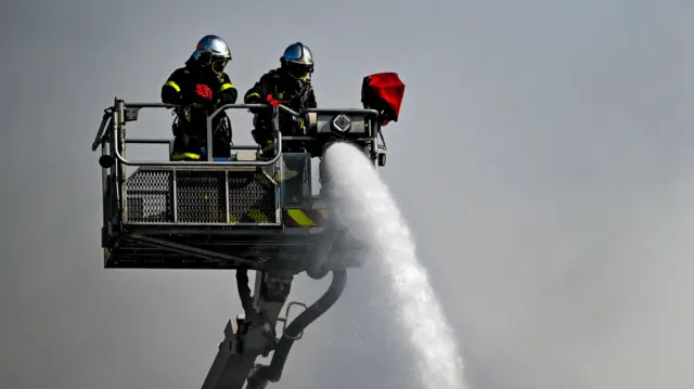 Dwóch strażaków na platformie z wężem strażackim i drabiną z koszem ratunkowym