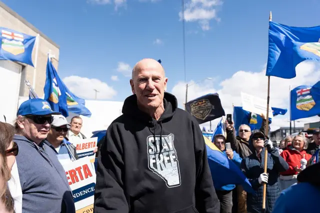 Mitch Sylvestre, director de Stay Free Alberta, un hombre de mediana edad, con poco pelo, viste una sudadera negra que lee "Queda libre" sobre el mapa de la provincia de Alberta. Está rodeado de simpatizantes que ondean banderas de Alberta