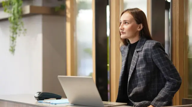 Kobieta z biura zaufania siedzi przy ladzie recepcyjnej i pracuje z laptopem - zdjęcie stockowe