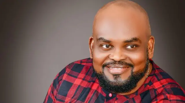 Oloogbe Solomon Akiyesi, o n rẹrin-in muse. O wọṣo alawọ pupa to ni dudu tẹẹrẹ lara.