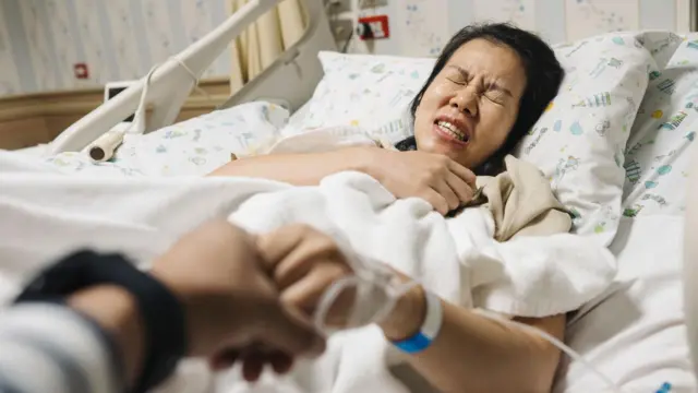 Una mujer en cama con una fuerte expresión de dolor toma la mano de otra mujer que está tomando la foto.