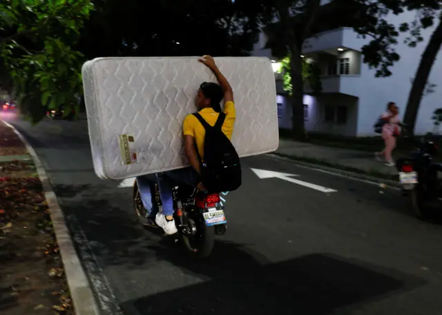 Dos personas viajan en una moto cargando un colchón entre ambas. 