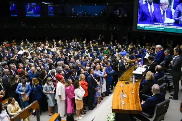 Lula toma posse no Plenário da Câmara dos Deputados. Lula lê um papel da mesa da Câmara enquanto uma multidão de pessoas em roupas sociais o observa do plenário
