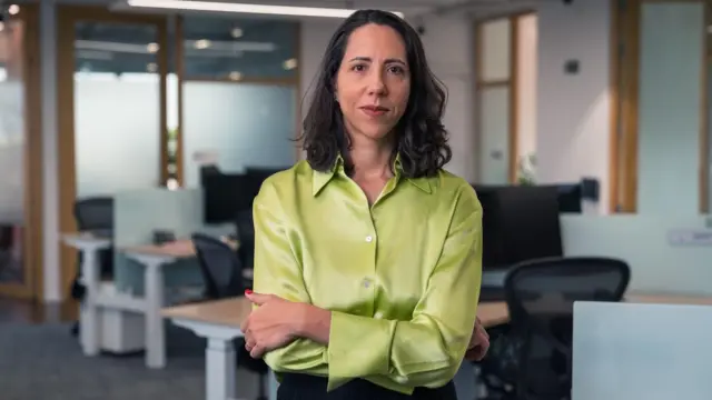 Laura Carvalho veste uma blusa verde de cetim e mangas longas, e tem os braços cruzados. Ela olha para a câmera com um sorriso discreto
