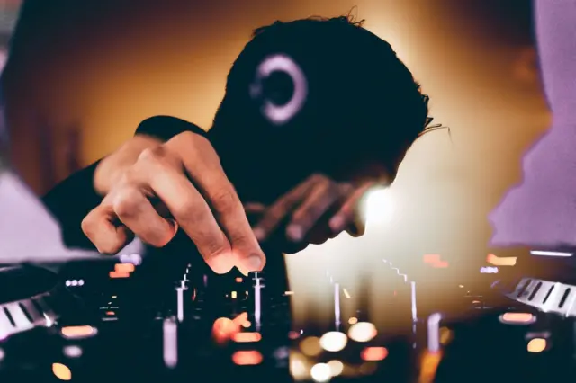 Крупним планом видно руку, яка керує музичним пультом: пальці налаштовують регулятори на мікшерному пульті. На тлі — яскраві розмиті вогні та клубна атмосфера.