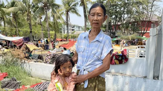 An elderly woman stands with her hands on the shoulders of a young girl near some makeshift tents