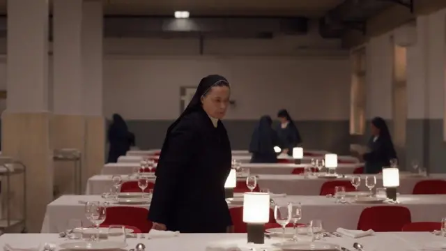 Монахиня стоит возле сервированного стола, на котором еще нет еды. На заднем плане видны еще столы и еще монахини.
