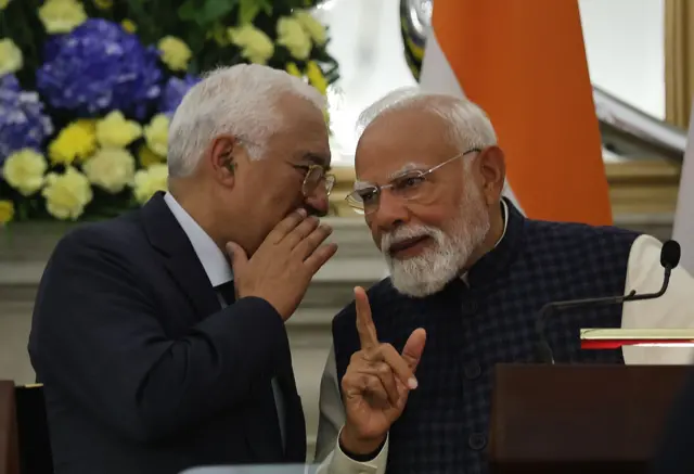 Кошта та Моді розмовляють пошепки. Кошта схилився ближче, прикриваючи рукою рот, наче передає конфіденційну репліку. Моді уважно слухає, піднявши вказівний палець, ніби роблячи акцент або збираючись відповісти. Позаду видно великі букети жовтих і синіх квітів та прапор Індії.