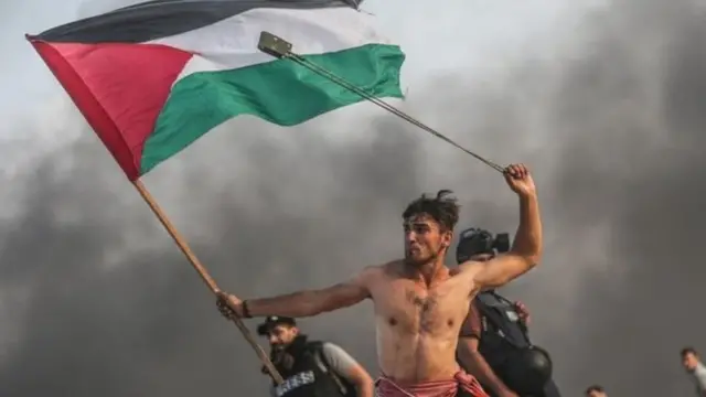 شاب عاري الصدر يحمل العلم الفلسطيني بيمينه ومقلاعا في يساره. صورة بعدسة الصحفي المصور محمد حسونة