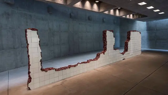 Uma estrutura revestida de azulejos brancos corta o centro de um espaço de concreto aparente, como uma parede fragmentada ou aberta à força. As bordas irregulares revelam camadas avermelhadas e orgânicas que lembram carne exposta, criando um contraste intenso entre a geometria limpa dos azulejos e a sensação de violência da obra.