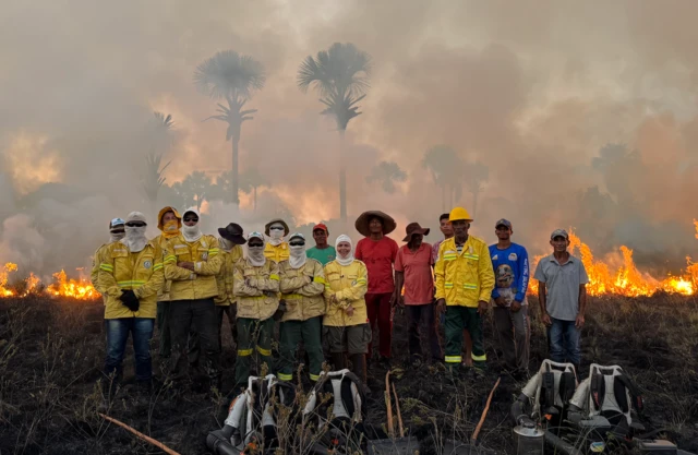 Vários brigadistas alinhados, alguns com jaquetas amarelas e os rostos cobertos com balaclavas, posam com chamas consumindo vegetação de Cerrado ao fundo