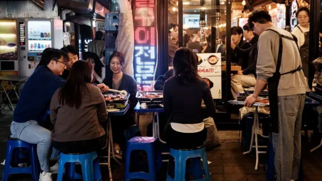 People eat barbecue on tables and stools outside a restaurant.