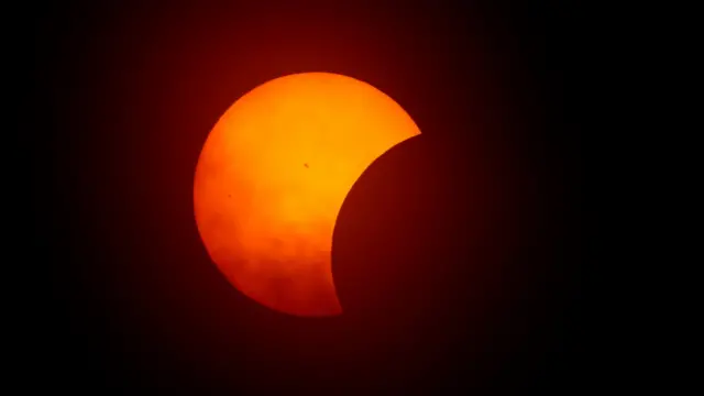 Foto colorida mostra o início do eclipse, com o Sol na partefoguetinho da betanotrás, laranja, e a lua o encobrindo como uma sombra