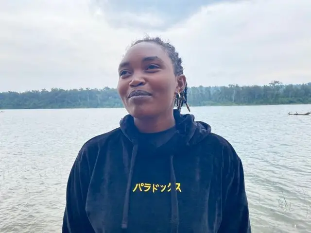 Umukobwa ureba hejuru gato ya 'camera', yambaye umupira uriho ingofero w'ubururu bwijimye ushushanyijeho mu gatuza n'indodo z'ibara ry'umuhondo. Imbere yambayemo undi mupira wijimye. Umusatsi we uboshye inweri. Inyuma ye hari ikiyaga, abantu bari mu bwato n'ishyamba hakurya y'ikiyaga. 