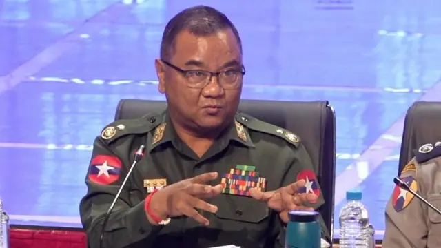 Thiếu tướng Zaw Min Tun trong quân phục đang ngồi tại một cuộc họp báo với ánh đèn màu hồng phía sau. Ông dang rộng hai tay khi đang phát biểu.