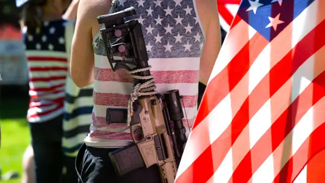 Hombre usa una camisa con la bandera de Estados Unidos y un rifle colgado en su espalda