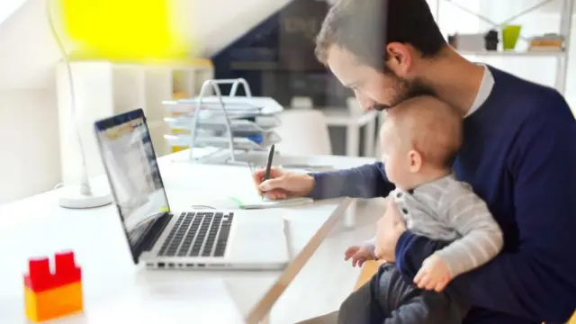 Seorang pria bekerja di depan laptop sambil memangku bayi.