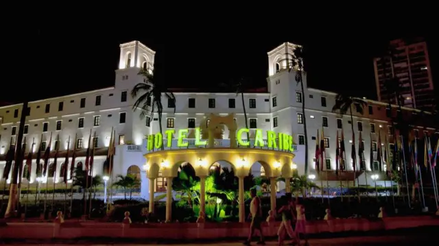 Una vista nocturna del Hotel Caribe de Cartagena, de arquitectura al estilo colonial, rodeado de palmeras y banderas de muchas naciones, iluminado con luces blancas. El nombre del hotel resalta en grandes letras de luz verde.