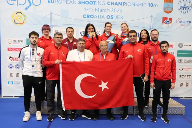 Milli takım türk bayrağı ile selam veriyor. Ortalarında Şevval İlayda Tarhan altın madalyasını ısırarak poz veriyor.