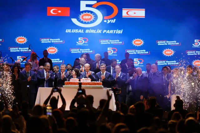 UBP logosu ve bayrakların olduğu bir fonun önünde kalabalık pasta kesiyor, alkış tutuyor.