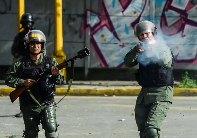 Agentes de la Guardia Nacional Bolivariana vestidos de verde y usando cascos disparan.