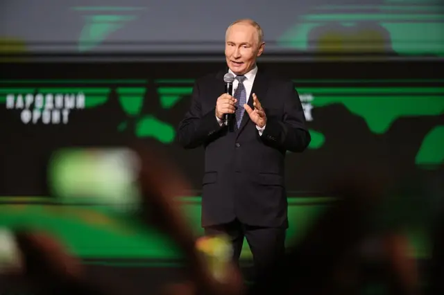 Қора костюмдаги Путин қўлида микрофон ушлаб саҳанада нутқ сўзламоқда.
