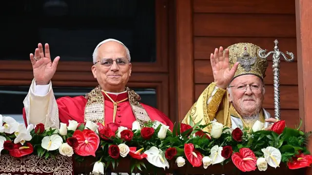 Papa Leo kırmızı tören kıyafeti, sağındaki Bartholomeos ise sarı renkli tören kıyafetiyle ellerini kaldırmışlar ve selamlıyorlar