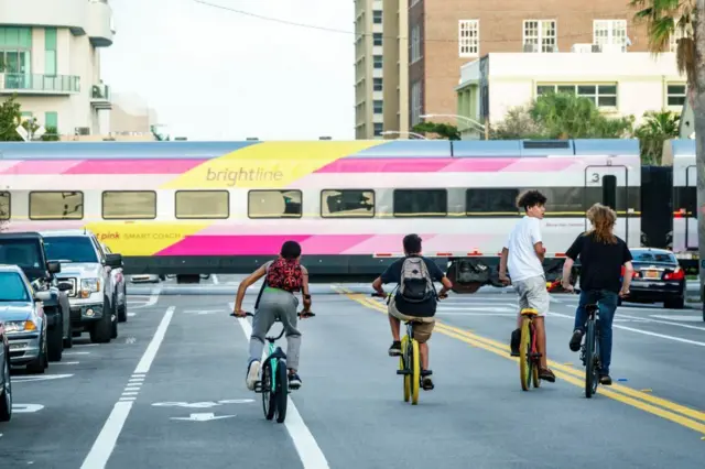 Cuatro niños montan en bicicleta por una calle por la que cruza un tren Brightline. 