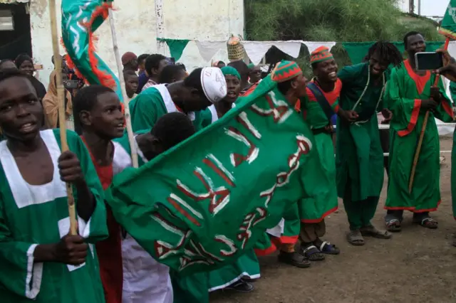 رجال صوفيون سودانيون يؤدون رقصة تقليدية خلال مهرجان ذكرى المولد النبوي في ولاية القضارف، بينما يرتدون جلاليب خضراء ويحملون يافطة  حضراء من القماش كُتب عليها "لا إله إلا الله، محمد رسول الله.