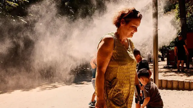 黄色いタンクトップを着た濃い色の髪のショートヘアの女性が、ミストの立ち込める公園を歩いている。周囲では子供たちが遊んでいる