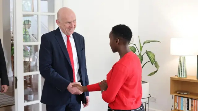 John Healy wearing a dark suit and red tie shaking hands with Esther Njoki, who hass a red jacket. A man's arm in a suit is visible through an open door begind Healy. 