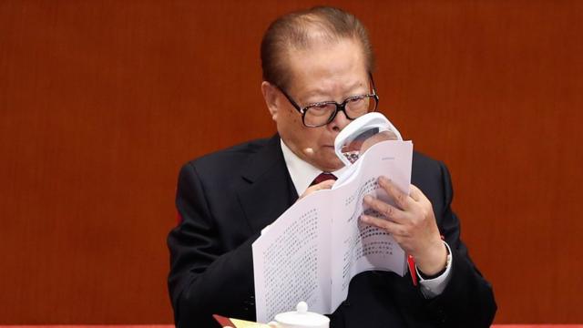 Jiang Zemin reads a document with the aid of a magnifying glass, while also wearing his trademark large spectacles.