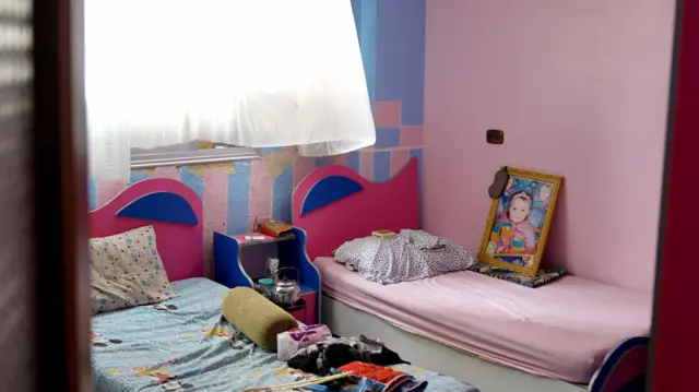 Mavi ve pembe duvarlar, iki yatak ve küçük bir kızın büyük çerçeveli bir resminin bulunduğu bir çocuk yatak odası görülüyor
